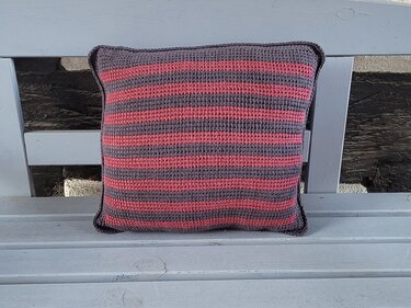 Quadratisches gehäkeltes Kissen mit horizontalen rosa und grauen Streifen auf einer hellgrauen Holzbank