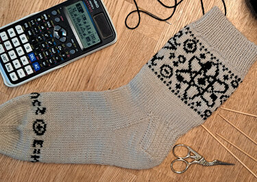 Beige handgestrickte Socke mit schwarzem Fair-Isle-Muster, Stricknadeln, Garn, Schere und Taschenrechner auf Holz.