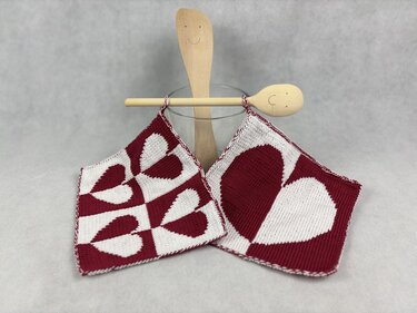 Two square knitted potholders in burgundy and white with heart patterns hanging from wooden spoons
