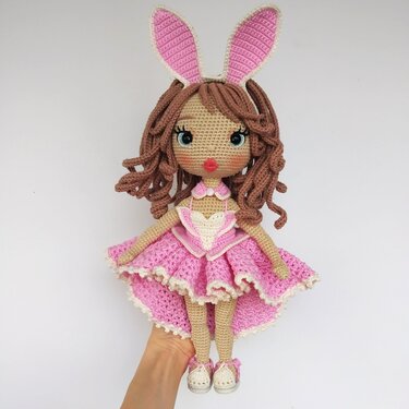 Handcrafted crochet amigurumi doll with curly brown hair, large eyes, pink high-low ruffled dress and pink bunny ears, held in a hand.
