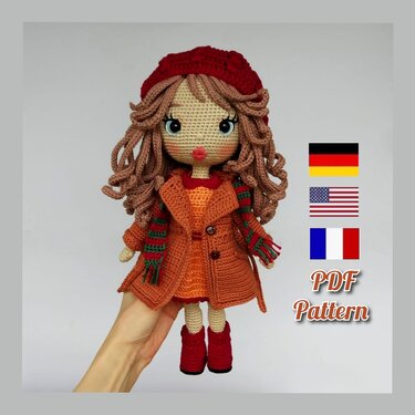 Hand holding a crocheted amigurumi doll with curly brown hair, red hat, orange coat, striped green-and-red scarf and red boots