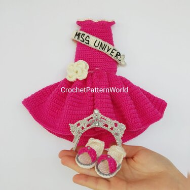 Bright pink crocheted doll dress with a cream sash and flower, a small decorative tiara, and two tiny pink-and-cream crocheted sandals held in a hand.
