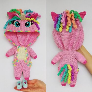 Front and back views of a pink crocheted unicorn doll jumpsuit with hood, colorful curly mane, small horn, tail, and a pair of light-blue shoes, held by a hand.