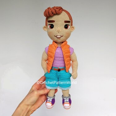 Crocheted amigurumi boy doll wearing orange vest, purple top, teal shorts, and two-tone sneakers, held by a hand