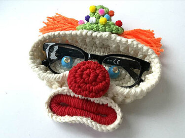 Gehäkelter Clown-Brillenhalter mit orangefarbenem Haar, roter Nase und grünem Hütchen, darin eine schwarze Brille.