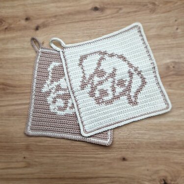 Zwei gehäkelte quadratische Topflappen in Creme und Beige mit Hundekopfmotiv auf Holzuntergrund.