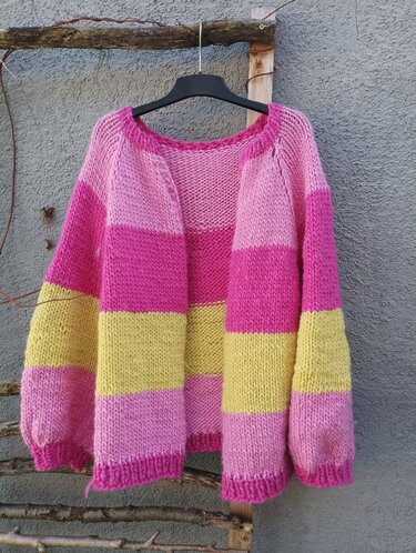 Grobgestrickte Damenjacke mit breiten pinken und gelben Streifen an einem schwarzen Bügel vor grauer Wand