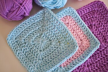 Close-up of three crocheted squares in light blue, pink, and purple with yarn balls.