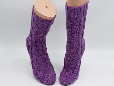 Gestrickte lila Socken mit Herz- und Zopfmuster auf Schaufensterpuppenfüßen vor hellem Hintergrund.