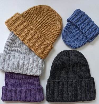 Five ribbed knitted beanies in camel, light grey, purple, charcoal, and a small blue cap on a light surface.