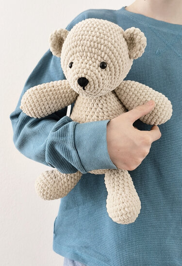 Person hält einen cremefarbenen, gehäkelten Amigurumi-Teddybären aus dickem, flauschigem Garn im Arm.