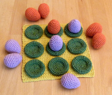 Gehäkeltes gelbes Spielbrett mit neun grünen Mulden und orangefarbenen sowie lila gehäkelten Eiern.
