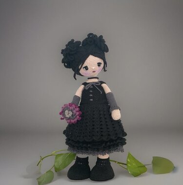 Crocheted amigurumi doll wearing a black dress, grey fingerless gloves, and black crocheted shoes, holding a small purple and grey flower bouquet.