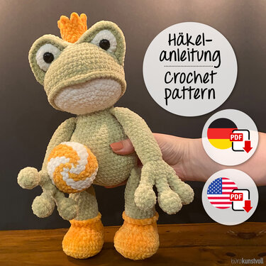 Crocheted frog amigurumi in pale green with an orange crown and accents, held by a hand; round labels indicate PDF downloads in German and English.