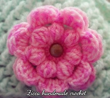 Nahaufnahme einer pinken gehäkelten Blume mit kleinem braunen Knopf auf hellblauem Häkelgrund