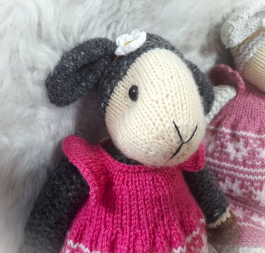 Gestricktes Amigurumi-Lamm mit rosa Kleid, grauen Ohren und Blüte im Haar auf hellem Hintergrund