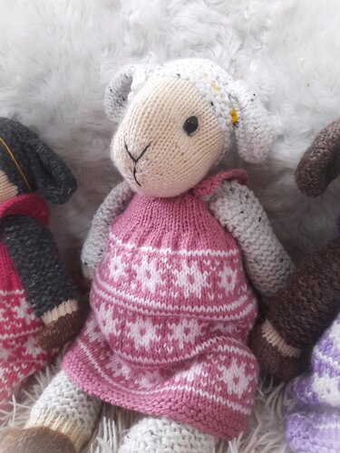 Gestricktes Amigurumi-Lamm mit beigem Kopf, schwarzen Augen und rosa-weiß gemustertem Kleid auf flauschigem Hintergrund