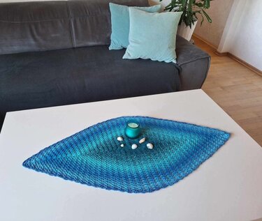 Gehäkeltes diamantförmiges Deckchen in Blautönen auf weißem Couchtisch mit Kerze und Muscheln