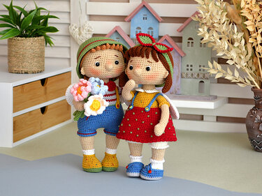 Two crocheted amigurumi dolls, one holding flowers, standing on a tabletop with decorative houses in the background