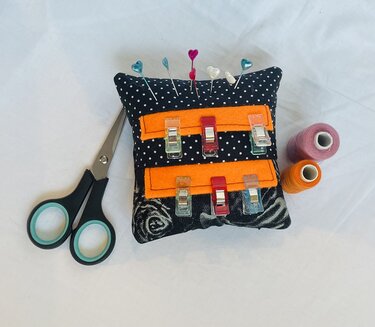 Nadelkissen aus gepunktetem Stoff mit orangefarbenen Filzstreifen, Stoffklammern, herzförmigen Stecknadeln, Schere und Garnrollen