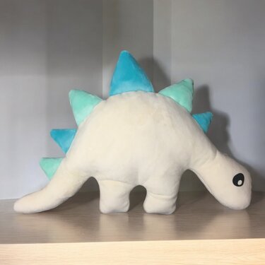 Cremeweißes Dinosaurier-Kuscheltier mit türkisfarbenen Rückenzacken auf einem Holzregal