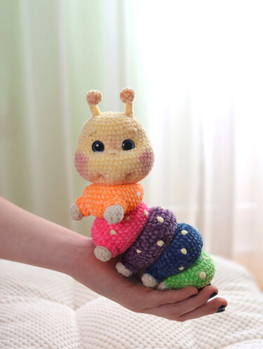 Gehäkelte bunte Amigurumi-Raupe mit gelbem Kopf und fünf farbigen Segmenten auf einer Hand