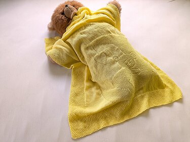 Teddy bear partially covered by a bright yellow knitted baby blanket with heart and bottle motifs.