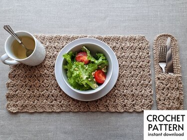 Top-down view of a crocheted placemat and matching cutlery holder in natural beige yarn with a salad bowl and mug on a light grey tablecloth.