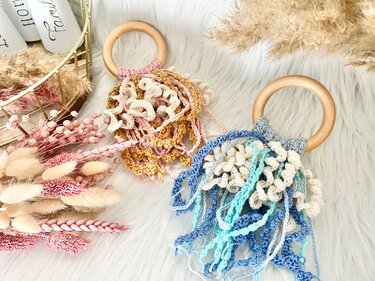 Zwei gehäkelte Greifringe mit Holzringen und gekräuselten Fransen, einer rosé-creme, der andere blau-creme, auf flauschigem Untergrund.