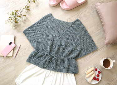 Overhead view of a dusty blue crocheted kimono-style top with a V-neck and drawstring waist laid on a light wood floor with lifestyle accessories.