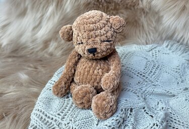 Gehäkelter brauner Amigurumi‑Teddybär aus Chenille‑Garn mit gestickten Augen auf heller gestrickter Decke