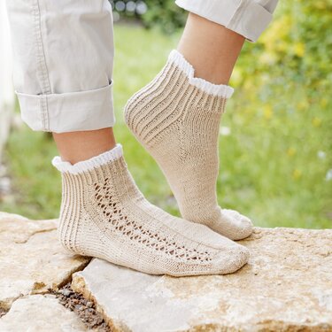 Beigefarbene, gestrickte Damensocken mit Lochmuster und weißem Rüschenrand an den Füßen einer Person auf einem Steinuntergrund.