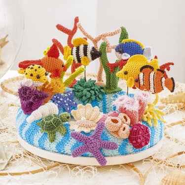 Gehäkeltes Korallenriff mit Fischen, Schildkröte, Seesternen und Muscheln auf blau-weiß gestreiftem Sockel.