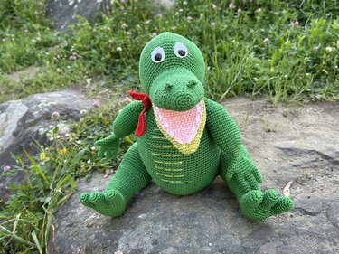 Grüner gehäkelter Amigurumi-Dinosaurier mit rotem Halstuch und geöffnetem Maul auf einem Stein im Garten