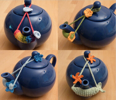 Blaue Teekanne mit vier unterschiedlichen gehäkelten Teebeutelhaltern an Ausguss und Deckelknauf