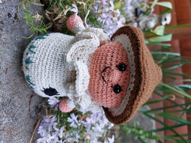 Gehäkeltes Amigurumi-Glückspilz mit braunem Hut, rosa Gesicht und weißem Rüschenkragen vor Blumen