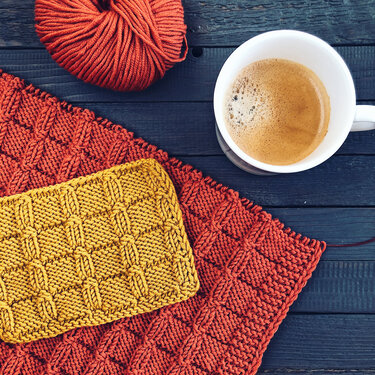 Close-up of textured square stitch swatch on rust blanket with yarn ball and coffee cup
