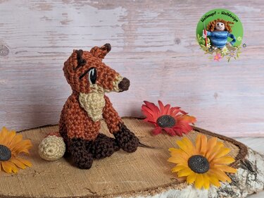 crocheted amigurumi fox in rust, cream and dark brown yarn seated on a wooden slice with artificial sunflowers