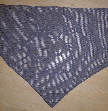 Gehäkeltes dreieckiges Tuch in Dunkelgrau mit eingearbeitetem figürlichem Motiv, liegend auf Holzboden.