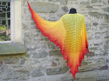 Triangular knitted lace shawl on a mannequin, colors fading from yellow through orange to red, against a rustic stone wall.