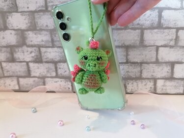 Green crocheted amigurumi dragon phone charm with pink accents on a light green phone, white brick background and pastel beads.