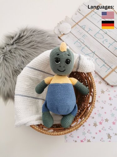 Crocheted green and blue amigurumi dinosaur with yellow horn sitting in a wicker basket on a white knit blanket