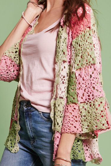 Damen trägt offene Häkeljacke aus Blumenquadraten in Rosa, Grün und Creme über T-Shirt und Jeans