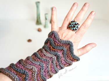 Hand wearing a multi-colored knitted fingerless cuff with wavy chevron ridges and a beaded ring.