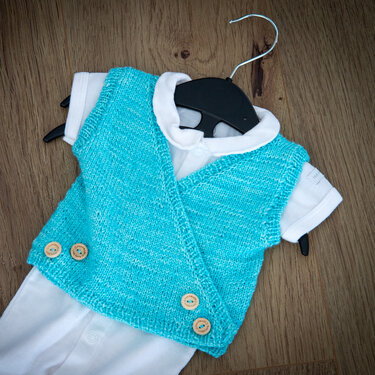 türkise gestrickte Wickelweste mit Holzknöpfen auf weißem Baby-Body auf einem Bügel liegend