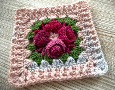 Handgehäkeltes Granny Square mit plastischen roten Rosen, grünen Blättern und zartrosa-grauem Rand