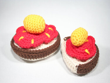 Nahaufnahme zweier gehäkelter Törtchen, ein ganzes und ein halbiertes, mit roter Blüte und gelber Kugel