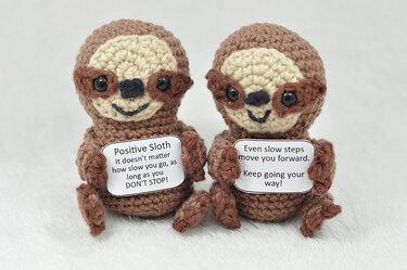 Zwei gehäkelte Amigurumi-Faultiere in Braun- und Beigetönen halten kleine Schilder mit aufmunternden Sprüchen.