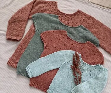 Vier gestrickte Baby- und Kinderpullover in Pastellfarben mit rundem Ausschnitt und punktartiger Struktur, nebeneinander auf hellem Stoff arrangiert.