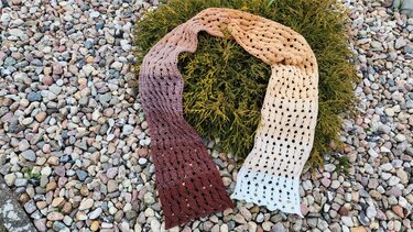 Long crocheted scarf with an ombre gradient from dark brown to cream, featuring an openwork lace pattern, laid on green foliage and pebbles.